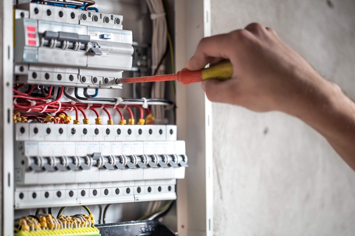 A hand working with a red-handled screwdriver on rows of circuit breakers in an open distribution board